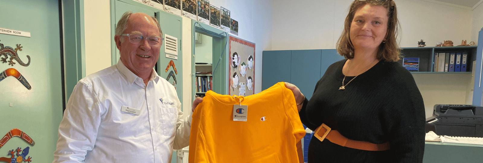 A man and woman standing in a school office, smiling and holding a donated yellow jumper betweenthem.