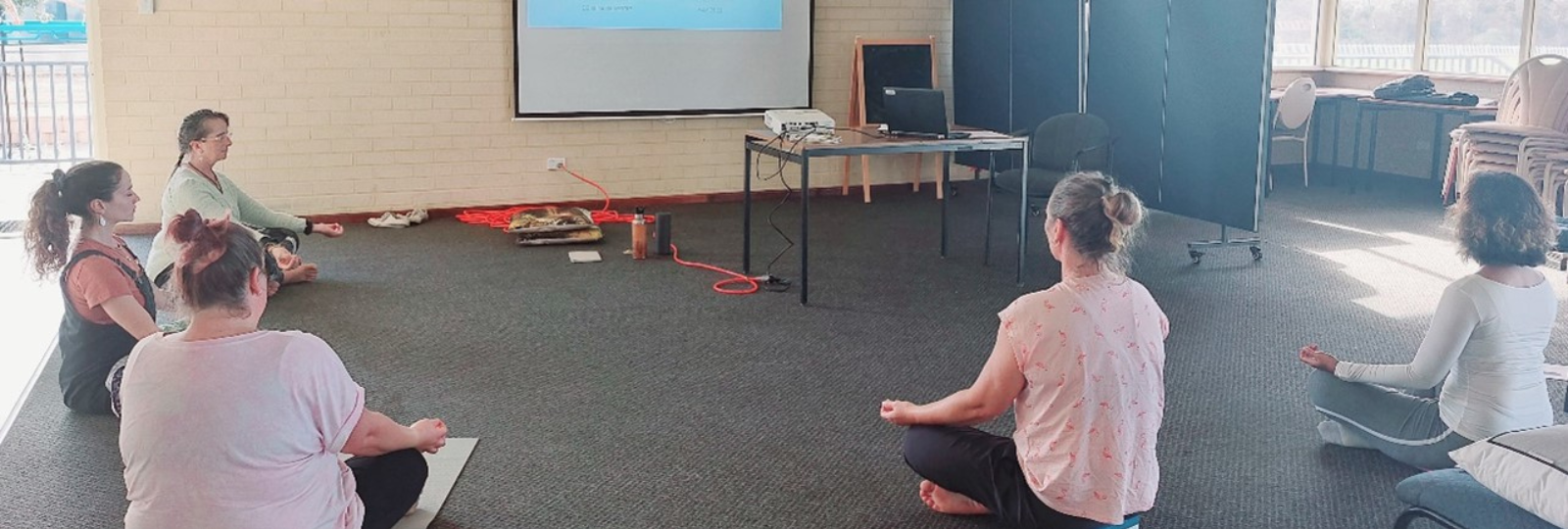 Five women seated in a semi circle in a room seated cross-legged during a yoga session