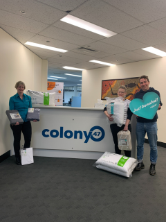 A male and female staff member from GIVIT, standing in a lobby with a female staff member from Colony47 holding donated linen.